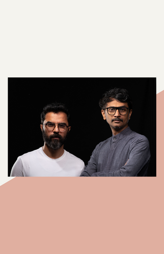 artists thukral and tagra standing next to each other against a black backdrop. one is wearing a white t-shirt and has a beard and glasses, the other wears a gray tunic and has glasses and a mustache